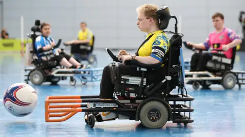The FA via Getty Images Picture of Dylan Kelsall during the match. He is sat in his wheelchair, with the ball ahead of him. He has blonde, curly hair and glasses.