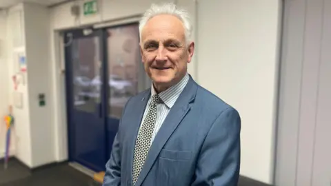 Man with grey hair in blue blazer stands in corridor 