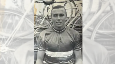 Reg Barnett A black and white photo of a young man staring at the camera with bicycles in the background