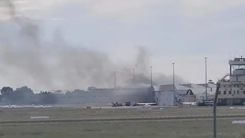 A far-away shot of smoke rising from an airport hangar in Adelaide after a plane crash. Aircraft can be seen stationed on the tarmac outside the hangar, with a green field in the foreground.