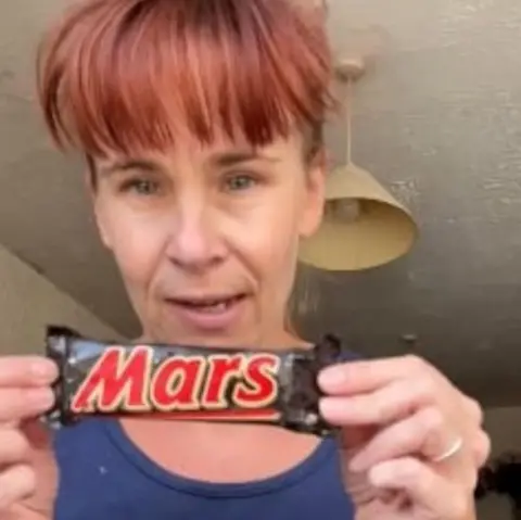 Victoria Gordon A woman holding up an old Mars Bar. She is wearing a navy vest and has red hair tied up with a full fringe.