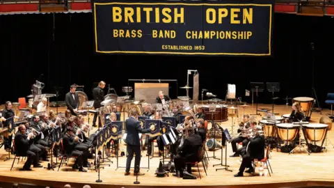 A brass band sat on a stage being conducted by a man stood up at the front. Behind is a large banner hanging on the wall that says British Open Brass Band Championships.