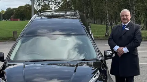 Moxon's Family Funerals Kevin Moxon, dressed smartly in uniform, stands by a funeral hearse. 