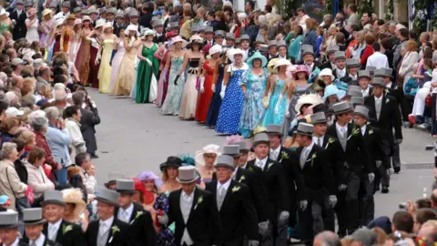 Helston Flora Day Association About 30 couples are walking along a street which is lined with spectators. The ladies are wearing brightly-coloured full-length gowns with hats and gloves. The gentlemen are wearing top hats and tails. The shops lining the street are decorated with greenery and flowers.