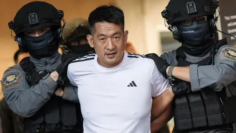 Getty Images A man in a white Adidas T-shirt being escorted by two guards wearing black helmets and protective vests. 