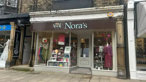 Julia Bryson/BBC A shop with books and clothes in the window. Above the window is a brown sign with blue writing that reads 'Nora's'.