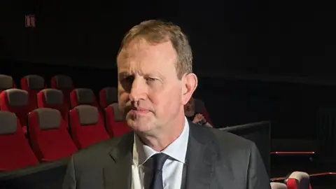 PA Jim O'Callaghan has fairish/brown short hair and is wearing a dark grey suit jacket, white shirt and dark tie. He is about to respond to a question put to him by a member of the media.