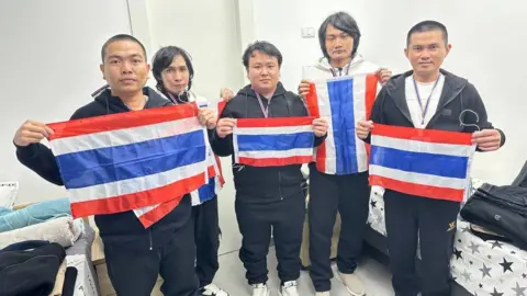 Royal Thai Embassy in Tel Aviv The five released hostages stand holding Thai flags in a white room. They are wearing tracksuits and trainers, with lanyards round their necks