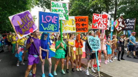 People dressed up in monochromatic colours of the rainbow holding up signs with their corresponding colours. The signs read phrases like "love wins" and "you are valid"