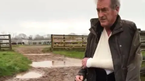 Jam Press Martin Dandridge in a brown jacket and white sling in a muddy field. 