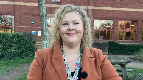 A woman with blonde curls and a light brown coat is sat outside the ambulance HQ on a picnic bench. She has her hands held together on the table and is smiling to the camera.