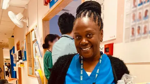 Supplied Nurse Neomi Bennett is in a hospital, wearing blue scrubs and a black cardigan. She is smiling and holding some equipment. 