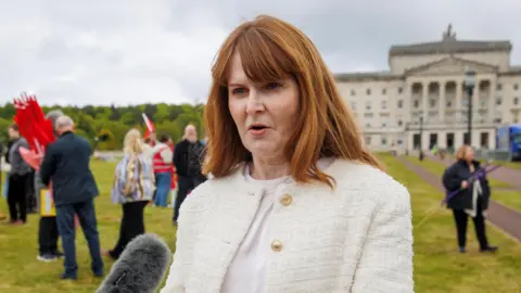 PA Media Caoimhe Archibald speaking into a microphone. She is wearing a cream jacket with gold buttons. She has long red hair. She is standing outside Stormont parliament buildings.