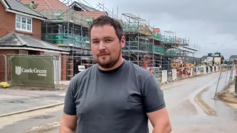 Nathan Long stand on the street in front of a houses with scaffolding and builder's fencing. 