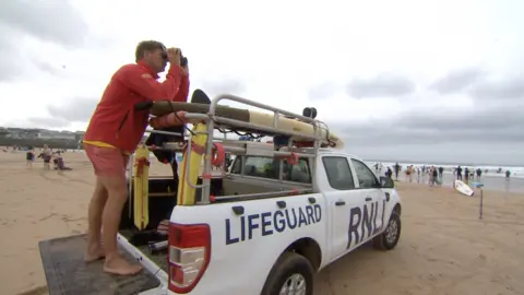 BBC A busy beach with choppy waves. An RNLI Lifeguard truck with a lifeguard standing on the back looking out to sea with binoculars. 