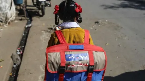 A view from the back of a young girl, dressed in a school uniform and carrying a school bag, walking on a street in India.