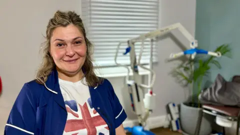 STEVE HUBBARD/BBC Oana Caspreac sits in he training room at Prime Care's headquarters. There is a hoist in the background to the left of her and a green plant. She is smiling at the camera wearing blue scrubs. Caspreac is wearing a white Union Jack T-shirt underneath the scrubs (which are unbuttoned). 