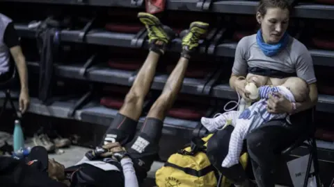Alexis Berg/Strava Sophie Power en una foto se sentó amamantando a su bebé. A la izquierda de ella hay un hombre que también es un corredor que yace con las piernas hacia arriba.