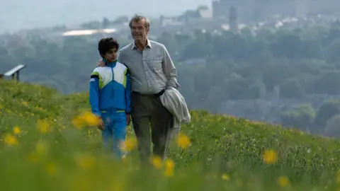Copyright of True Brit Entertainment An elderly man in a smart shirt strolls through a flowery meadow on a Summer's day. His arm is affectionately placed around the shoulders of a young boy dressed in a blue tracksuit