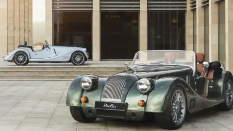 Morgan A green classic car, with an open top and a number plate which reads "PLUS SIX", sits in front of a building. Another classic car, a pale blue colour, is in the background.