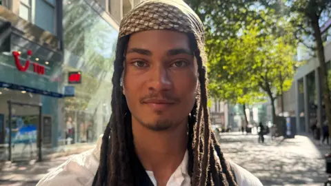 Leon has brown and black braided hair that sits past his shoulders. He wears a brown patterned bandana, white shirt and smiles at the camera