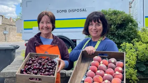Kim Hartshorne Two women - grocery shop workers, carrying trays of cherries and peaches while standing in front of a bomb disposal vehicle. Kim Hartshorne is not pictured - the women in the picture are her staff