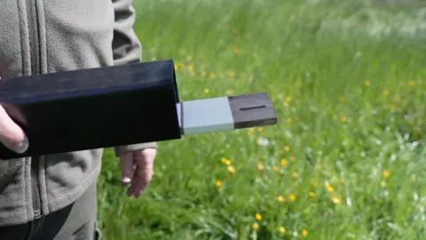 BBC A hollow black plastic rectangle with a narrow wooden board covered with white paper inside being held against a green field with yellow flowers