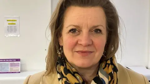 Woman with shoulder length fair hair looking at the camera, she is wearing a cream coat and has a black, mustard, white and grey scarf on.