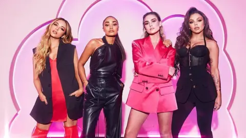The four members of Little Mix stand side by side wearing fashionable black and red fashion outfits in front of a pink backdrop