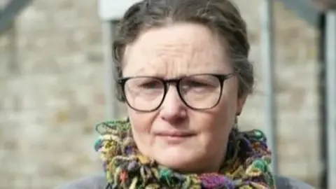 A headshot of Katie Sullivan. She is wearing a multi-coloured scarf and glasses and is unsmiling.