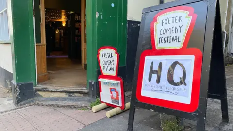 A sign with the words 'Exeter Comedy Festival HQ' written on it outside a pub