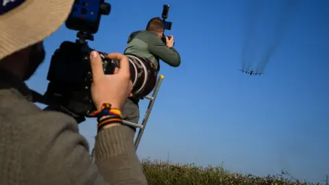 Two plane spotters taking a photograph of a United States Air Force B-52 Stratofortress as it takes off at Fairford RAF. They are wearing green coats and jumpers and there is a blue sky in the background.