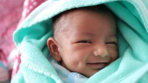 A close-up image of a baby's smiling face wrapped in a light blue hood