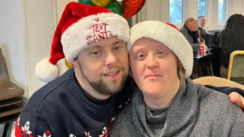 Michael Gallagher sits next to his girlfriend Alison Williams. He has his arm around her and their faces are together as they look into camera. Michael has a beard and Alison has shoulder length blond hair. They both look happy and are wearing Santa hats. Behind them is a room of people and some festive balloons as they're at a Christmas party. 