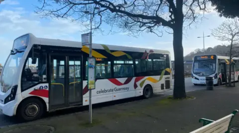A bus parked by a bus stop sign. It is multi-coloured and has the words 'celebrate Guernsey' written on the side.