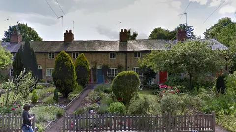 A terrace of brick cottages with different colour front doors, long adjoining front gardens full of trees and flowers, and a wooden fence across the front. A lady is walking past one of the two paths leading up to the cottages.