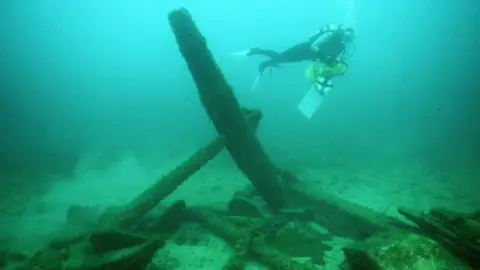 Valerie van Heest An underwater shipwreck, the water has shades of green and blue, a diver is swimming by what looks like an anchor