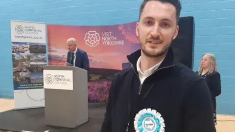 A bearded man in a black zipped top wearing a blue and white rosette looks at the camera