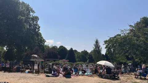 A crowd of people, mainly families, inside and surrounding the paddling pool. Some have parasols to protect from the sun. There is grass in the foreground and trees in distance