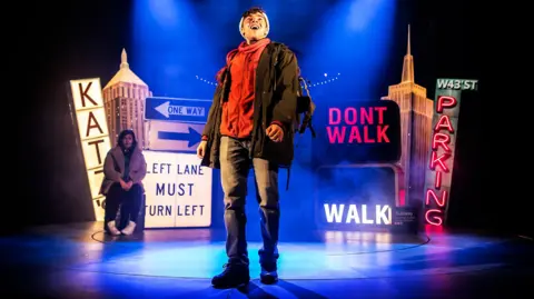 Pamela Raith Photography Two actors on a stage, the man at the front is wearing a hat, jeans, boots, a red jumper and a coat. A woman is sat behind him, looking at him, wearing white trainers, a coat and dark trousers. There are signs all around them. 