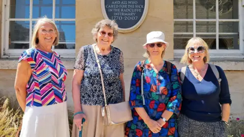 Four women smile and the camera in front of a plaque honouring the Radcliffe Nurses.