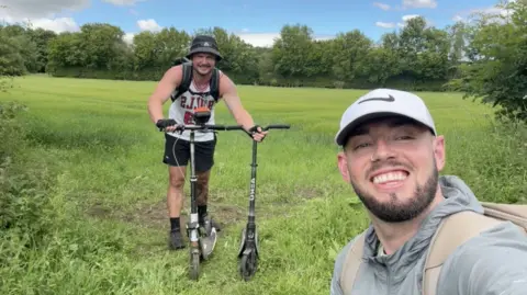 A man holding two black push scooters while standing in a field. He is wearing a backpack, a black bucket hat, a white vest, black shorts and trainers. In front of him taking the selfie is a man wearing a white Nike-tick hat and a grey coat, with a beige backpack. They are standingd in a field.