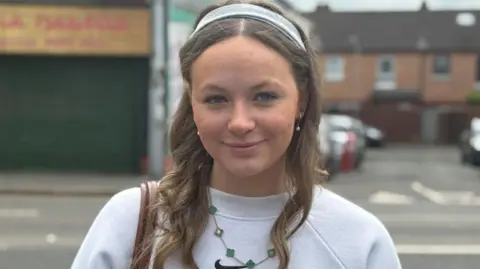 Naoise McCrory is wearing a grey headband and has her long, light coloured hair curled. She is smiling at the camera. On her right shoulder she is holding a brown leather bag strap and is wearing a grey crewneck jumper with a black Nike tick in the centre. She is stood in front of a closed shop front and an alleyway but the background is blurred.