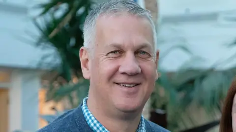 MSG Alan Rimington wearing a grey jumper over a white and light blue checked buttoned shirt. He is clean shaven and has short white to grey hair. He is smiling and stood in front of a tree that is slightly blurred.