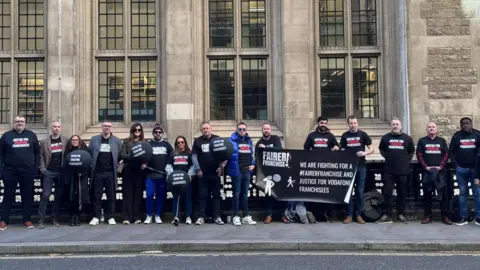 Fairer Franchise A group of people stand in a line outside a large stone and glass building. They are dressed mostly in black and holding black banners and balloons, all carrying a message about "Fairer Franchise". A banner reads: "We are fighting for a #FairerFranchise and justice for Vodafone franchisees".