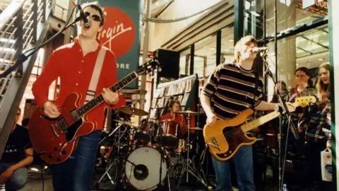 Getty Images A image of band performing in a shopping centre. The men play guitars in forefront while a man plays a drumset in the background. 