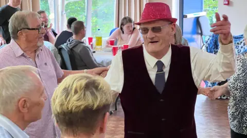 Pat dancing with a group of people - he holds his left hand aloft and is singing or speaking. Others stand in a circle around him holding hands, while there are tables in the background with guests sitting and drinking lemonade.