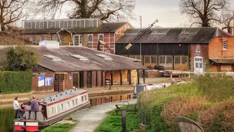 Ian Roberts A scenic shot of the yard and its surrounds with two people sat on a barge on the canal in front of buildings at the historic yard