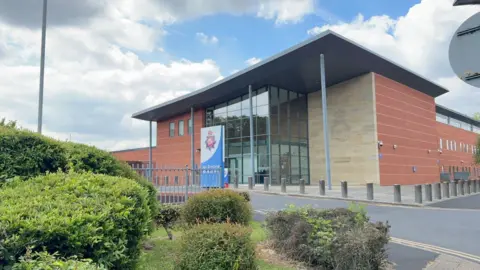 There is a glass foyer to the front of Bury Police Station. There is a sign in front of the building with the Greater Manchester Police logo.