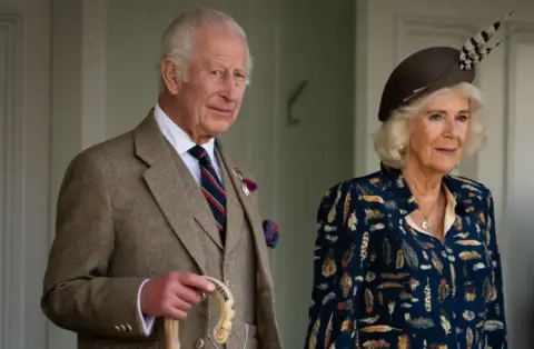 PA Media King Charles wearing a tweed suit with shirt and tie holding a shepherd's crook and standing next to Queen Camilla who is wearing a blue patterned dress and a blue hat with two feathers attached